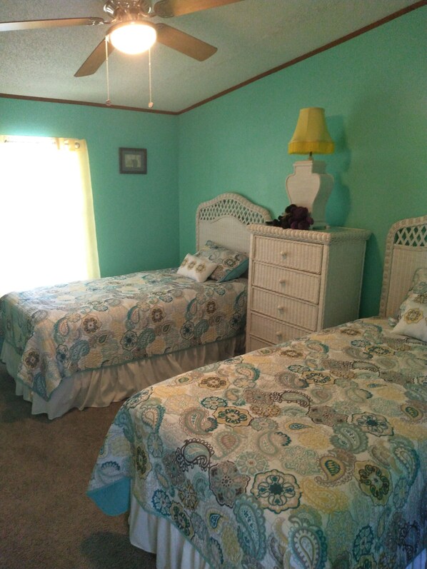 A bedroom with two single beds, a ceiling fan, a nightstand with a lamp, and a window with curtains. - "Serenity Now" Beautiful Lake Front property with Pool in Sunset Beach, NC (Sunset Beach)