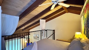 Un dormitorio con cama, ventilador de techo, ventana con cortinas y mesita de noche con lámpara.