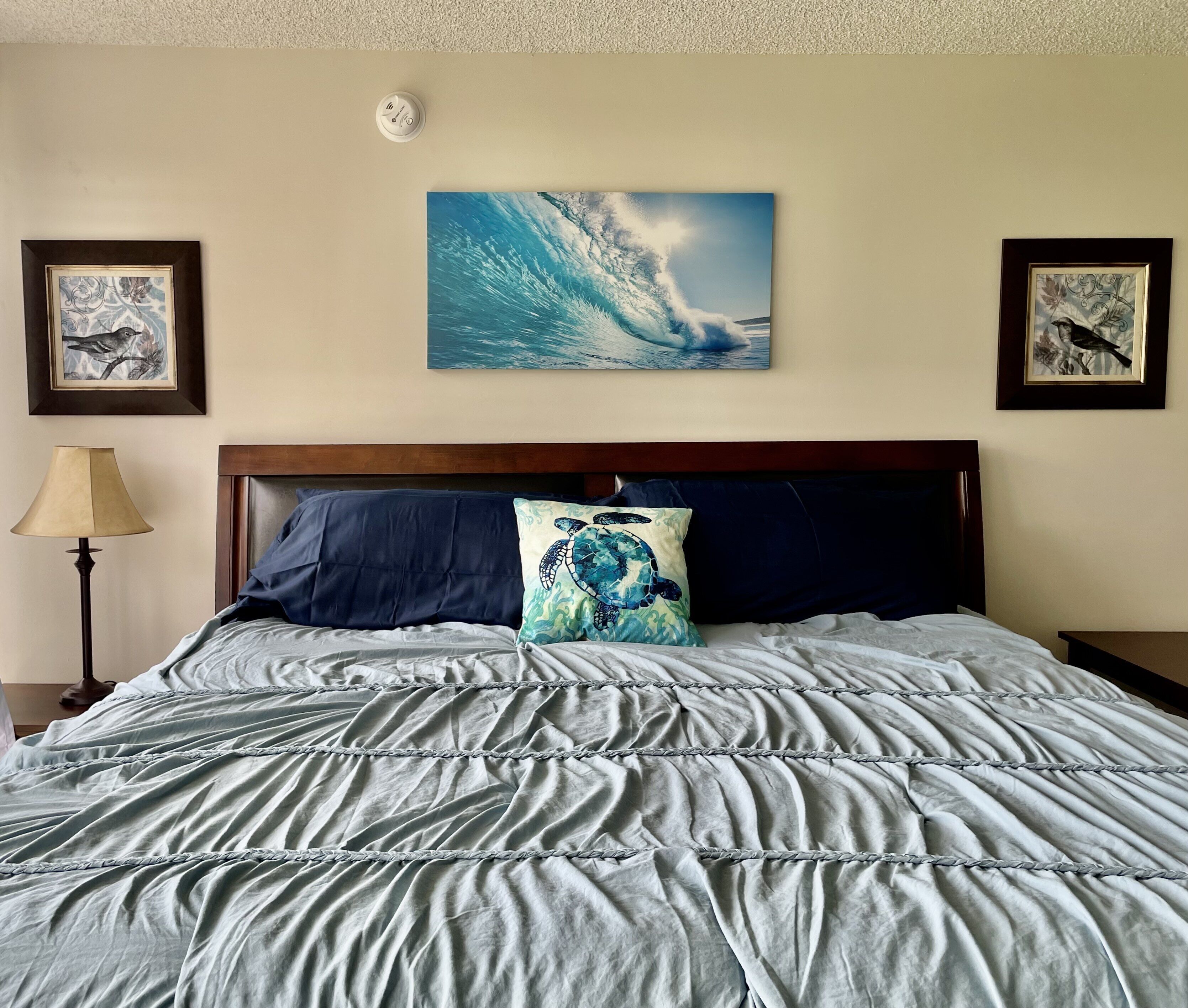 Una cama con colcha a rayas azul y blanco, un cojín con diseño de tortuga marina y un cuadro de una ola en la pared.