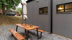 A wooden picnic table with benches, a metal umbrella, and a small window on a grey metal building. - Eva's Zen House | 2BR/2BA | Near Mercedes-Benz (Atlanta)