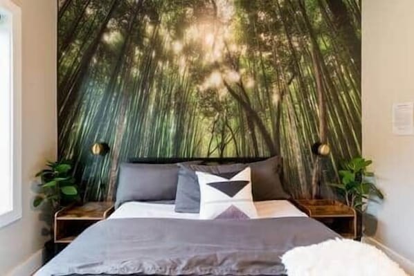 Un dormitorio con un gran mural temático de bosque, una cama con ropa de cama gris y blanca, y dos mesitas de noche con lámparas.