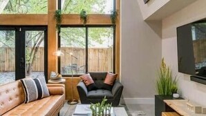 A modern living room with a leather sofa, a dark armchair, and a coffee table. There is a large window with a view of greenery outside. - Eva's Zen House | 2BR/2BA | Near Mercedes-Benz (Atlanta)