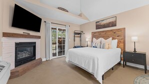 A bedroom with a fireplace, a bed with white bedding, a TV mounted on the wall, and a painting above the bed. - SADIE'S COTTAGE - Private Hot Tub and Fire Pit (Branson)