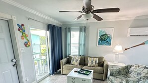 Una sala de estar con un ventilador de techo, un sofá, un sillón estampado, una mesa de centro y un cuadro en la pared.