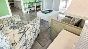 Una sala de estar con pared verde, un televisor de pantalla plana, una consola de madera, un sofá con estampado tropical y un sillón color beige.