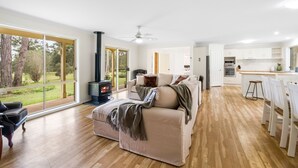 A spacious living room with a large window, a fireplace, a sofa, and a kitchen area with white cabinets and a wooden floor. - Bundara- Hunter Valley Retreat | Sleeps 11 | Rural | Resting amongst the trees (Congewai)
