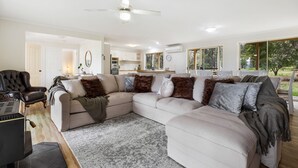 A spacious living room with a large sectional sofa, a coffee table, and a fireplace. There is a dining area with a table and chairs, and a kitchen in the background. - Bundara- Hunter Valley Retreat | Sleeps 11 | Rural | Resting amongst the trees (Congewai)