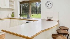 A modern kitchen with white cabinetry, a large island with stools, a window with a view of trees, and a wall clock. - Bundara- Hunter Valley Retreat | Sleeps 11 | Rural | Resting amongst the trees (Congewai)