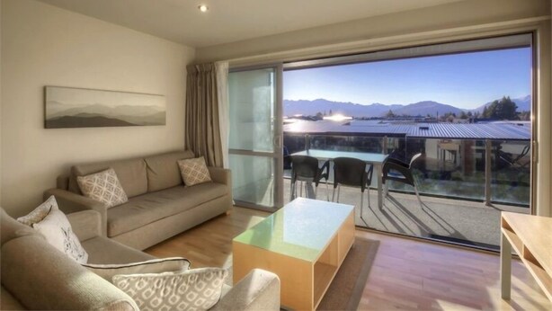 A modern living room with a sofa, armchairs, a glass coffee table, and a view of mountains through large windows. - Rocky Mountain Apartment - 111-112 (Wanaka)