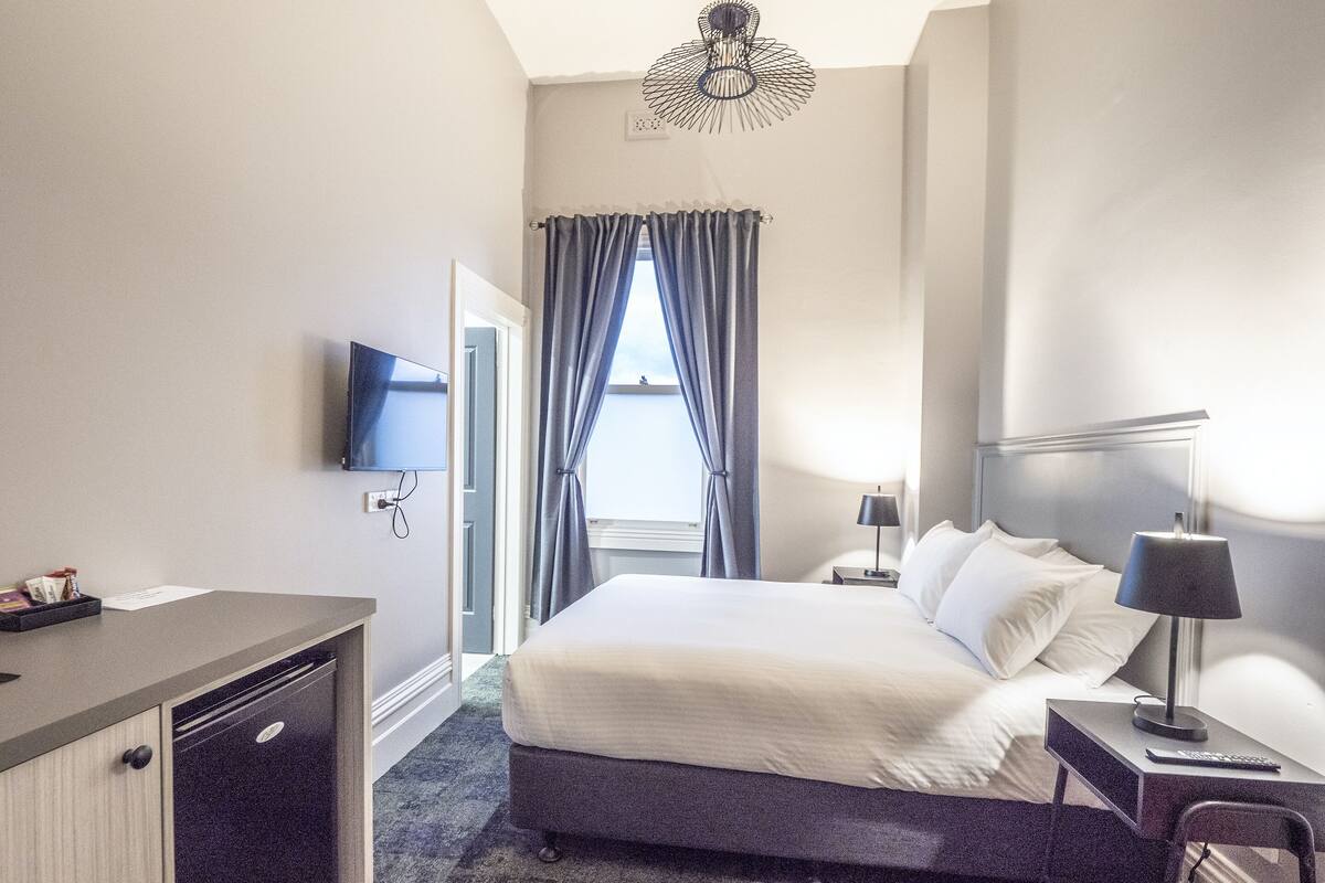 A hotel room with a bed, bedside table, lamp, TV, and a desk with a chair.