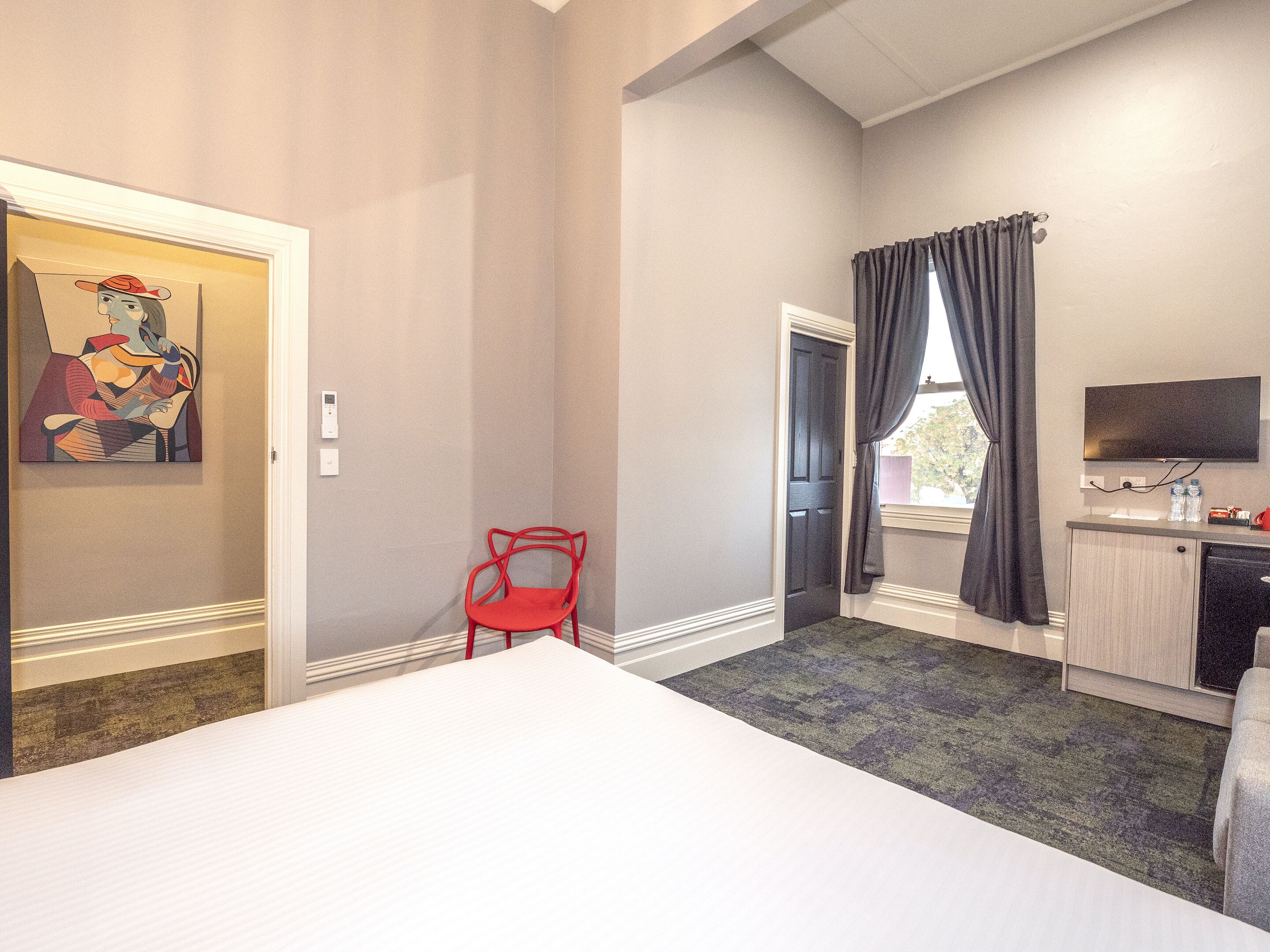 Habitación de hotel con una cama, un escritorio con televisor, una silla roja y un cuadro en la pared.