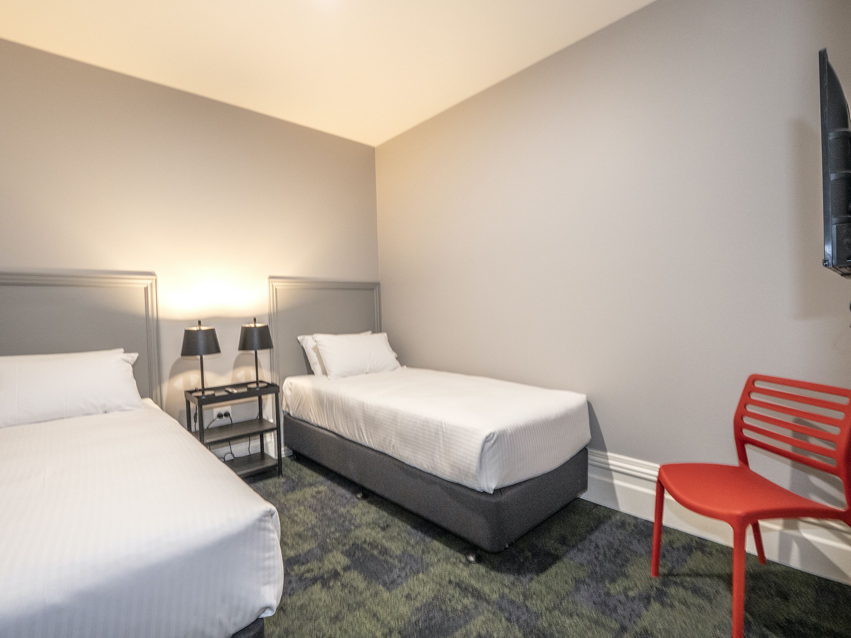 Habitación de hotel con dos camas, una mesita de noche con lámparas, una silla roja y un televisor montado en la pared.