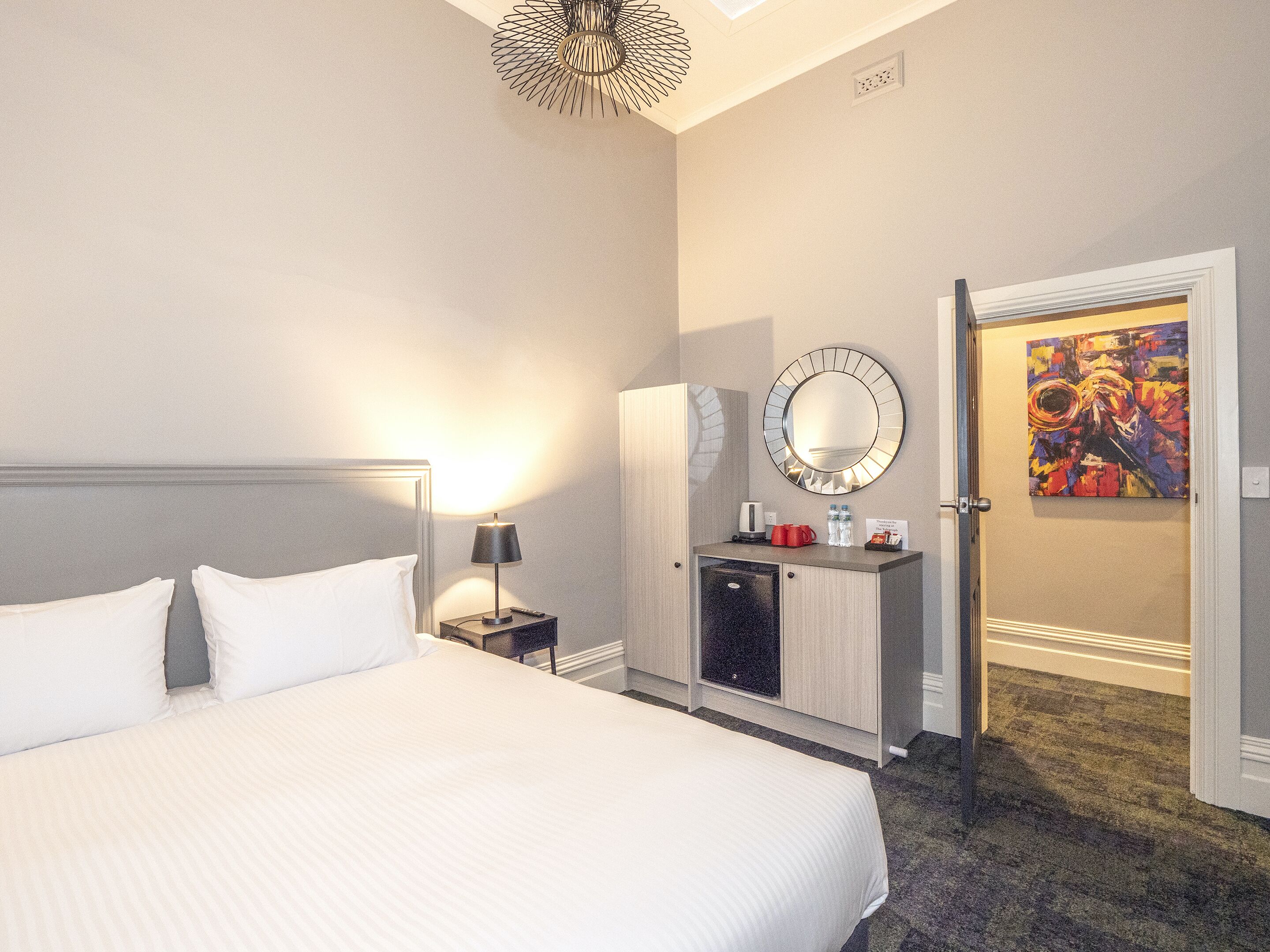 Una habitación de hotel con una cama grande, una mesita de noche con lámpara, un minibar, un espejo y un cuadro en la pared.