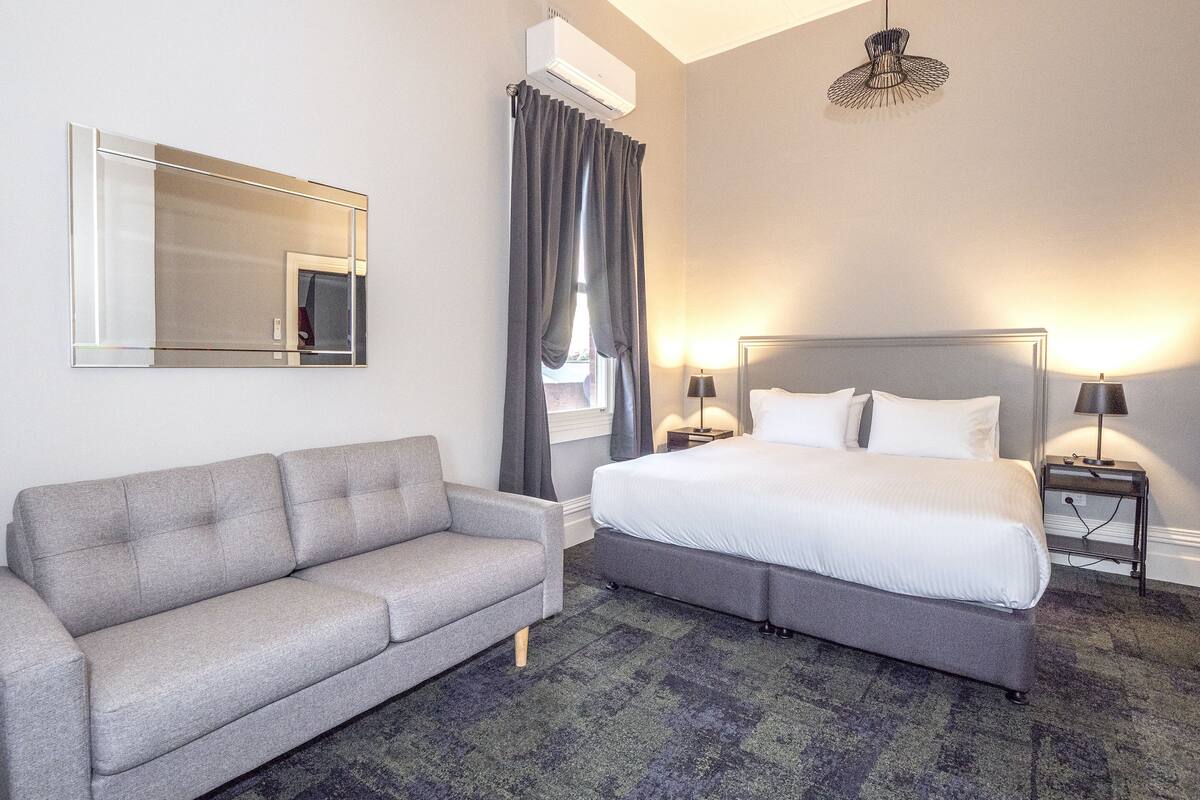A hotel room with a large bed, a grey sofa, a mirror, a window with curtains, and a ceiling light fixture.