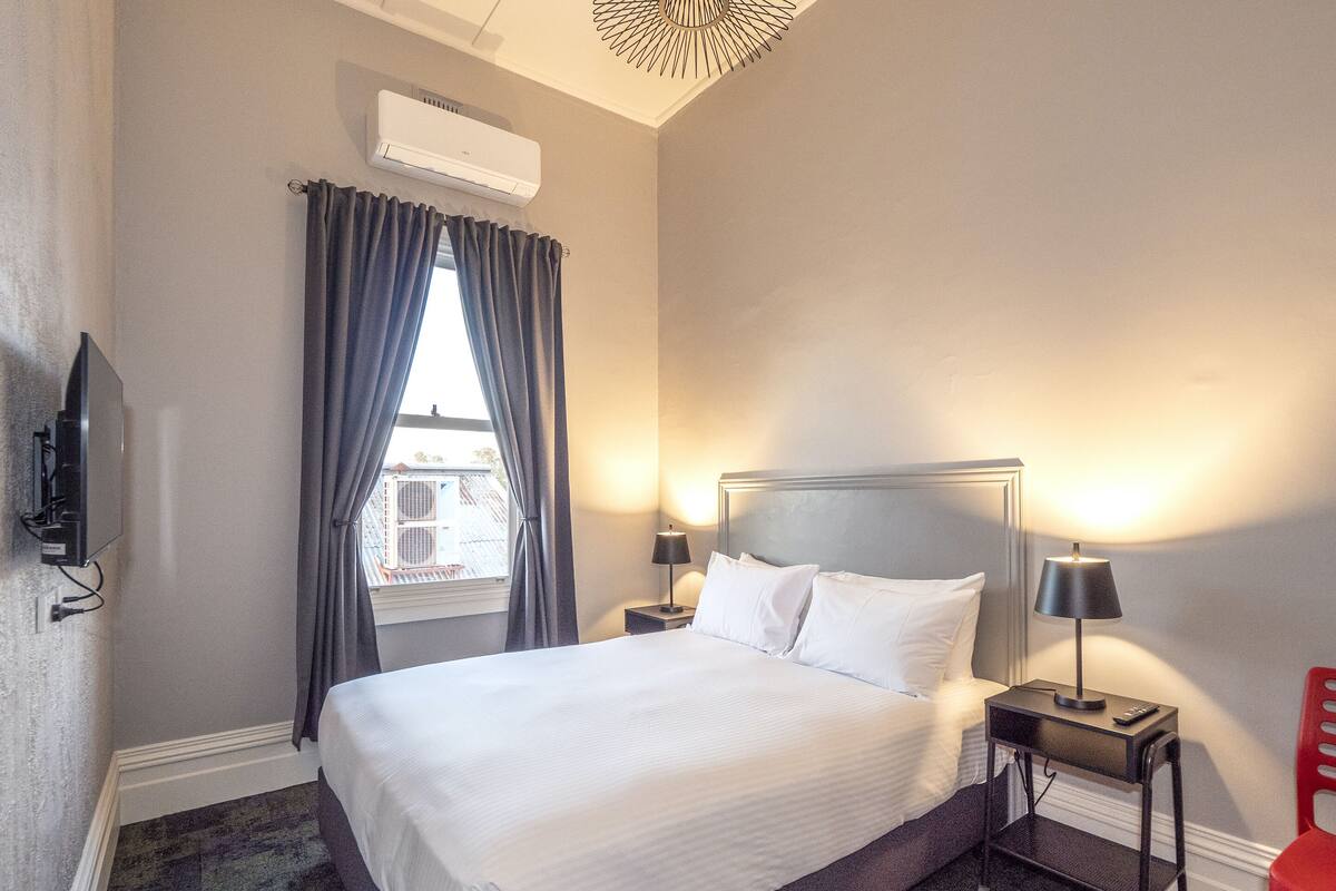 A hotel room with a bed, two bedside lamps, a TV, and a window with curtains.