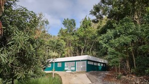 A small, single-story building with a green exterior, surrounded by dense vegetation and a gravel path leading to the entrance. - The Green House in Vega Alta, Zen zone close to the beach (Vega Alta)