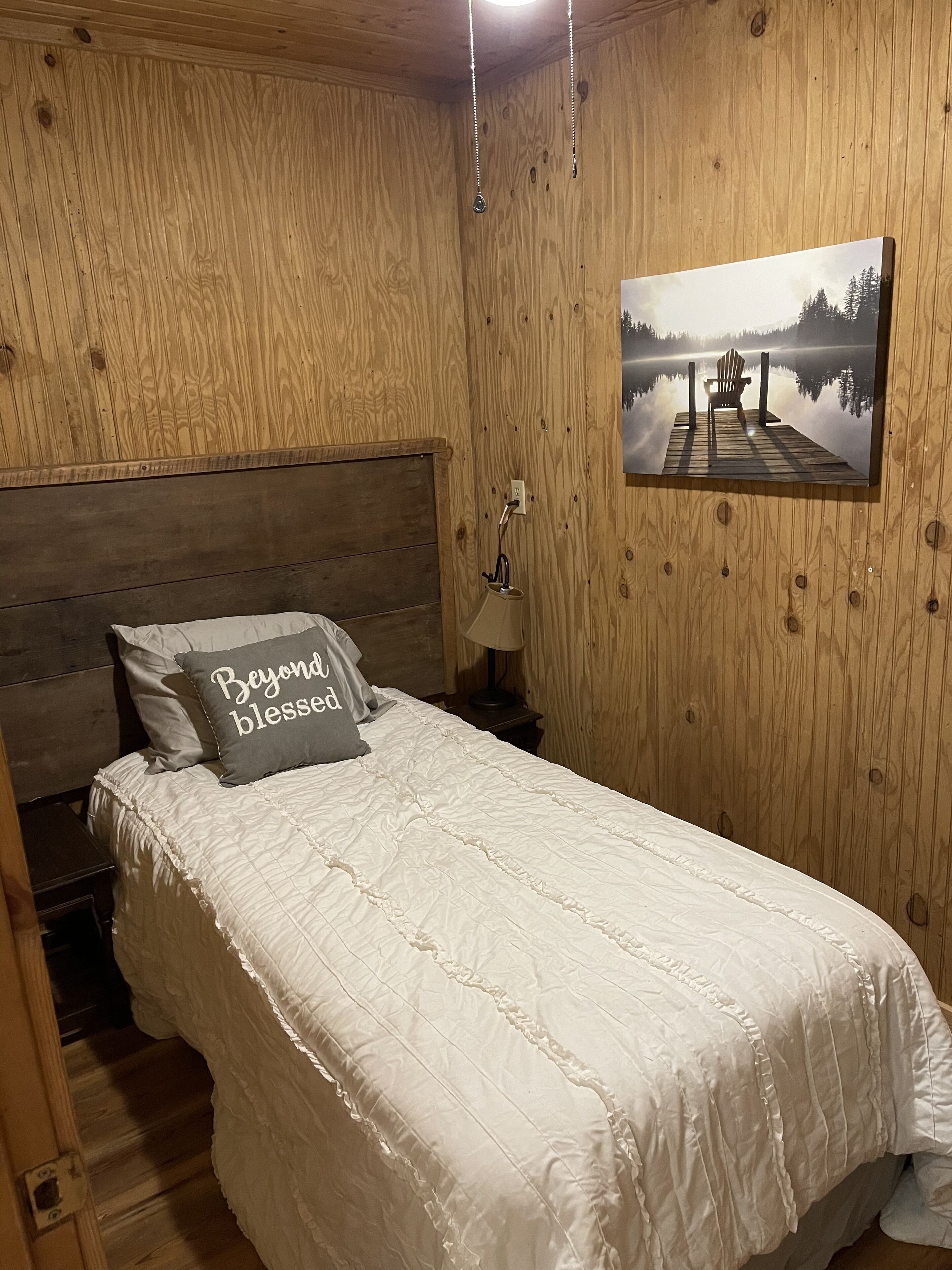 A wooden-paneled room with a bed, a nightstand, a lamp, and a picture of a dock with a chair.