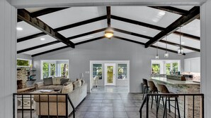 An open-plan living area with a high ceiling, exposed beams, a kitchen area with a bar, and a seating area with a sofa and chairs. - River View Retreat LLC (New Braunfels)