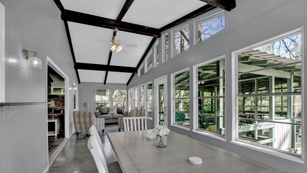 A spacious dining area with a long table, white benches, and large windows offering views of the outdoors. - River View Retreat LLC (New Braunfels)