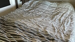 Un lit bien fait, avec une couvre-lit à rayures, une tête de lit sombre et un mur de couleur claire avec un revêtement texturé.