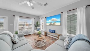 Ein Wohnzimmer mit großem Fernseher, Deckenventilator und Blick auf den Strand durch die Fenster.