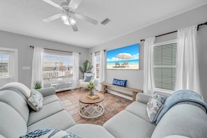 Una sala de estar con un televisor grande, un ventilador de techo y vista a la playa a través de las ventanas.