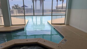 Ein Hallenbad mit Panoramafenstern, einem Whirlpool sowie Blick auf Palmen und einen Strand.