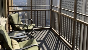 A balcony with two green cushioned chairs and a glass table, overlooking a cityscape with tall buildings.