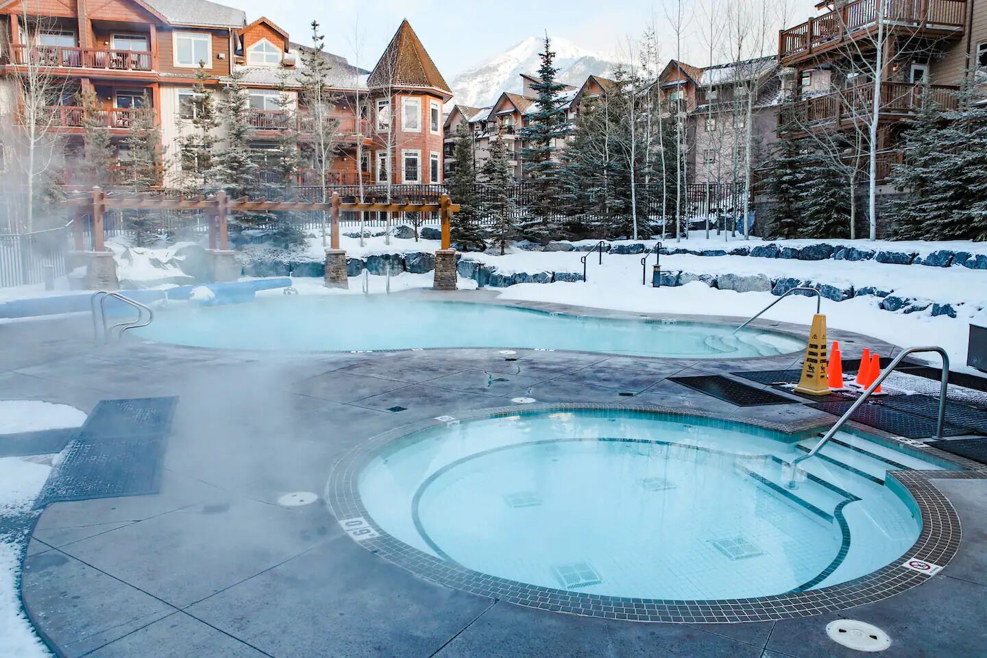 A hot tub area with a pool, surrounded by snow-covered ground and buildings with wooden balconies.