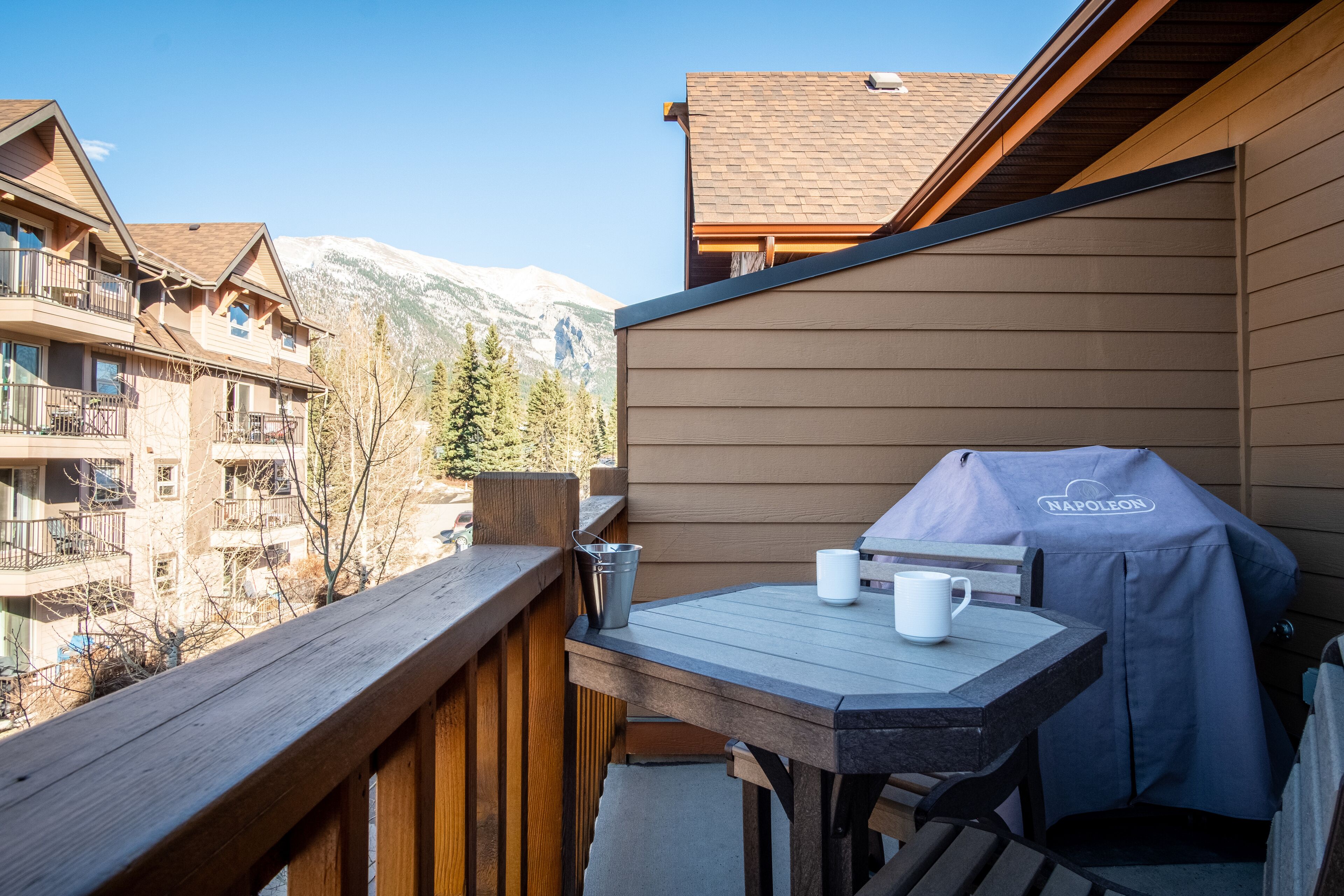 A balcony with a wooden railing, a table with a barbecue grill, and a view of snow-capped mountains.