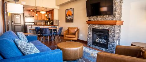 Una sala de estar moderna con un sofá azul, un sillón de cuero y un reposapiés, una chimenea de piedra y una cocina al fondo.