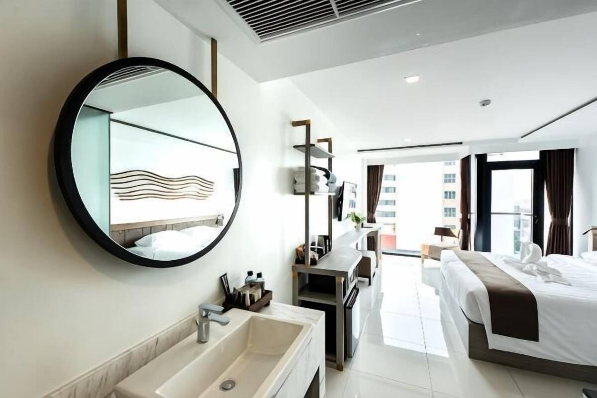 A modern hotel room with a large round mirror, a long sink, a bed with a white and brown bedspread, and a view of the city through large windows.