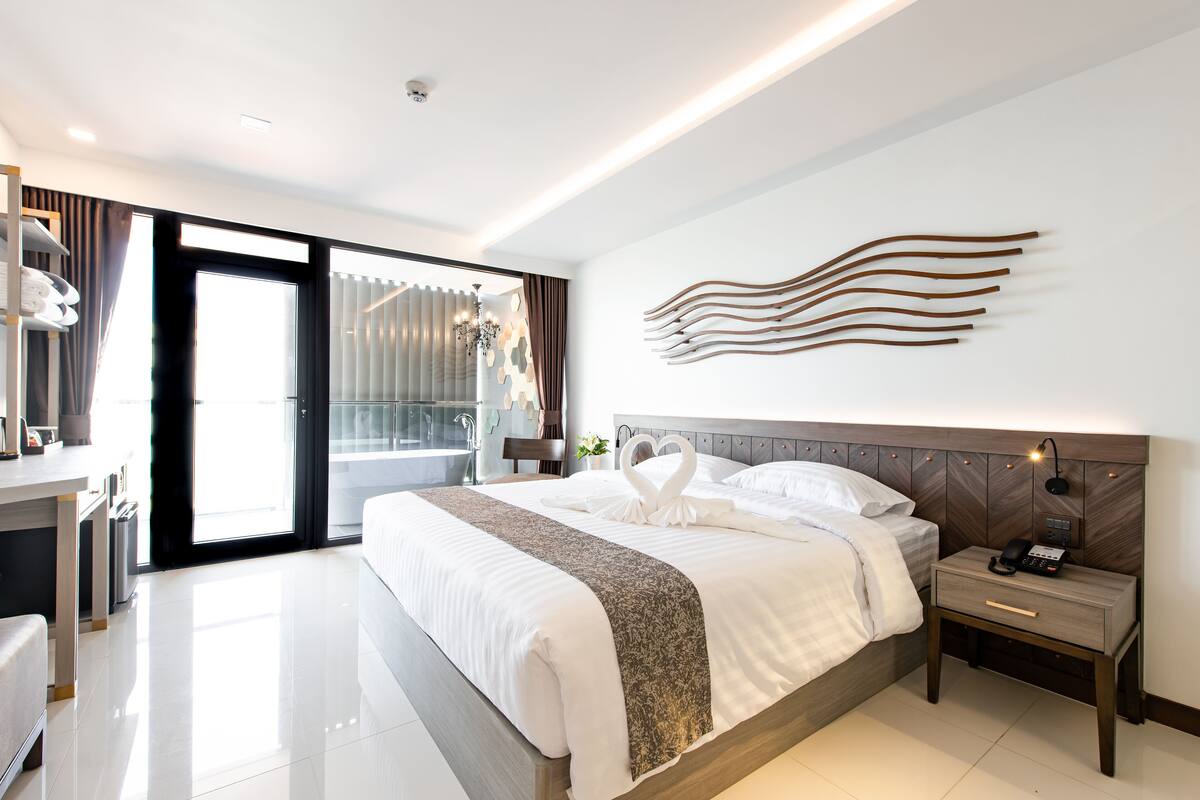 A modern hotel room with a large bed, a bedside table with a lamp, a wall-mounted artwork, and a sliding glass door leading to a balcony.