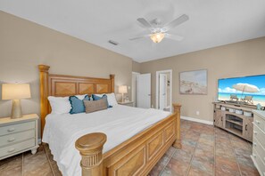 A bedroom with a large bed, a ceiling fan, a TV, and a beach-themed artwork.