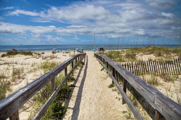 Un sentier qui longe la plage et mène à l’océan, avec une clôture en bois et des parasols de plage.