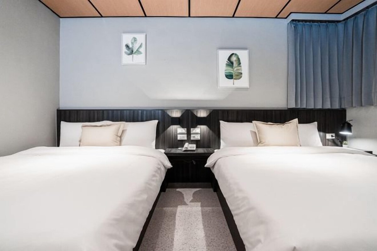 Una habitación de hotel con dos camas, un cabecero oscuro, una mesita de noche con lámpara y cuadros en la pared.