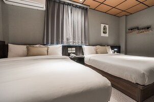 A hotel room with two beds, a window with curtains, a bedside table with a lamp, and a wall-mounted coat rack.
