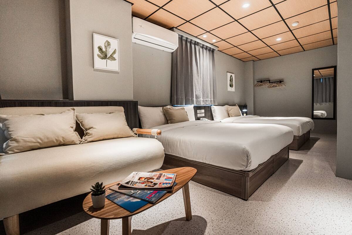 A hotel room with a large bed, a sofa, a small table with magazines, an air conditioner, and a mirror.