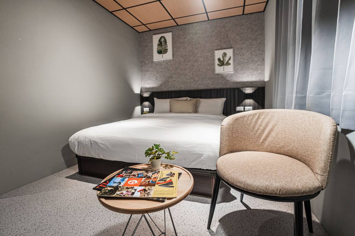 A hotel room with a neatly made bed, a small round table with a plant and magazines, and a comfortable armchair.