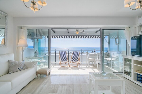 Ein modernes Wohnzimmer mit einem weißen Sofa, einem Glastisch, gestreiften Strandstühlen und Meerblick durch große Glasfenster.