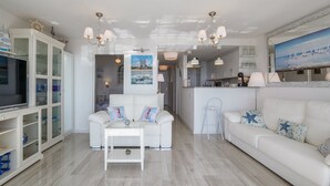 Un salon moderne avec un canapé blanc, une table basse blanche et un coin repas, la cuisine étant visible au fond.