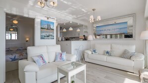 Un salon moderne avec un canapé blanc, une table basse en verre et un grand tableau représentant une scène de plage.