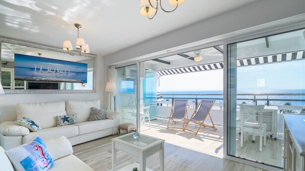 Un vaste espace de vie comprenant un canapé, une table basse et un tableau représentant des voiliers. Un balcon avec des chaises offre une vue sur la mer.