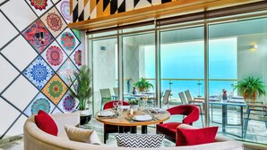 A modern dining area with a round table set for four, surrounded by red upholstered chairs. A large window offers a view of the ocean. - Tolip Resort Elgalala Hills (Ataqah)