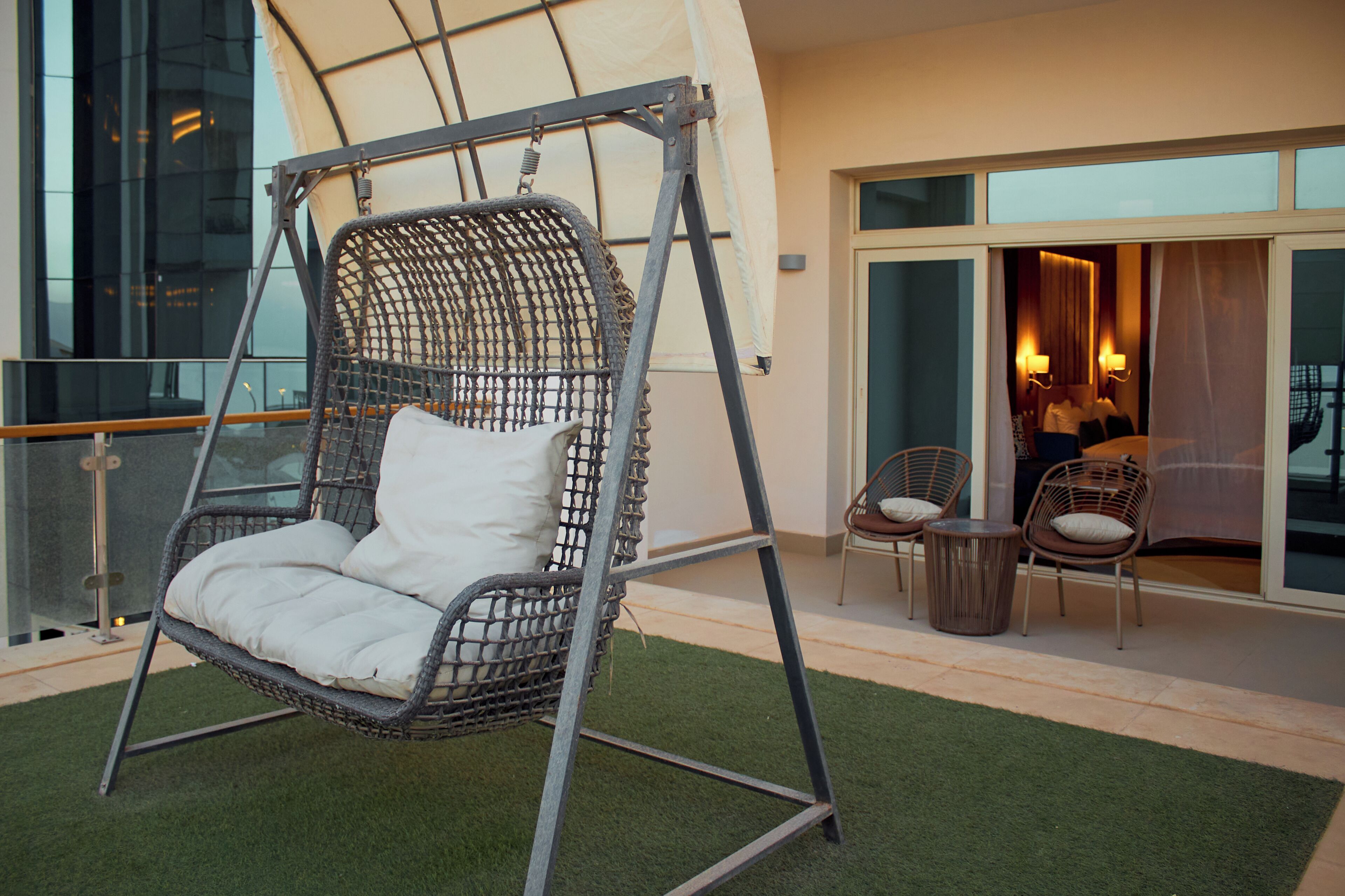 Une chaise suspendue avec des coussins, une petite table ronde et deux chaises avec coussins, le tout sur un balcon offrant une vue sur un immeuble.