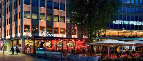 A multi-story building with a restaurant named 'Olivia' on the ground floor, outdoor seating, and a sign for 'ARNTZENDEBES' on the upper level.