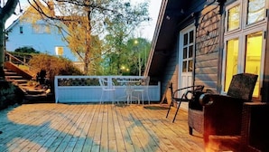 Una terraza de madera con mobiliario exterior, una parrilla y una casa al fondo.