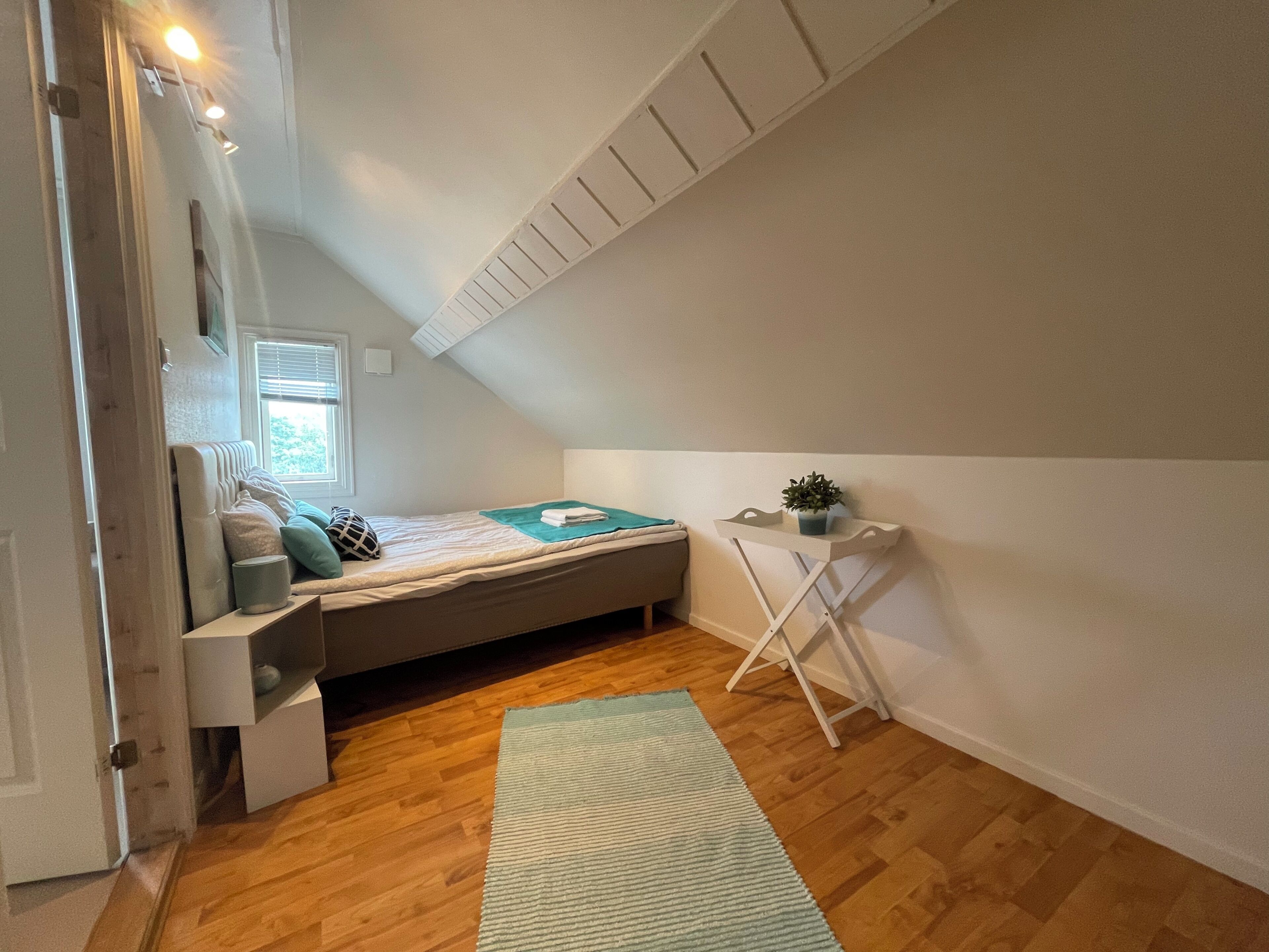 A small, cozy room with a sloped ceiling, a bed with pillows and a blanket, a small table with a plant, and a window with blinds.