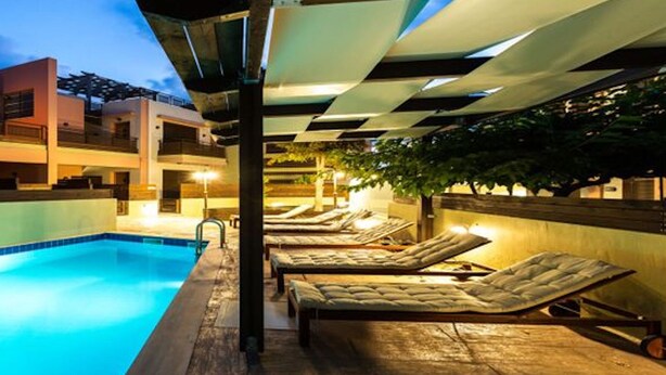 Una piscina con sillas de descanso y una zona cubierta, rodeada de edificios con balcones e iluminación exterior.