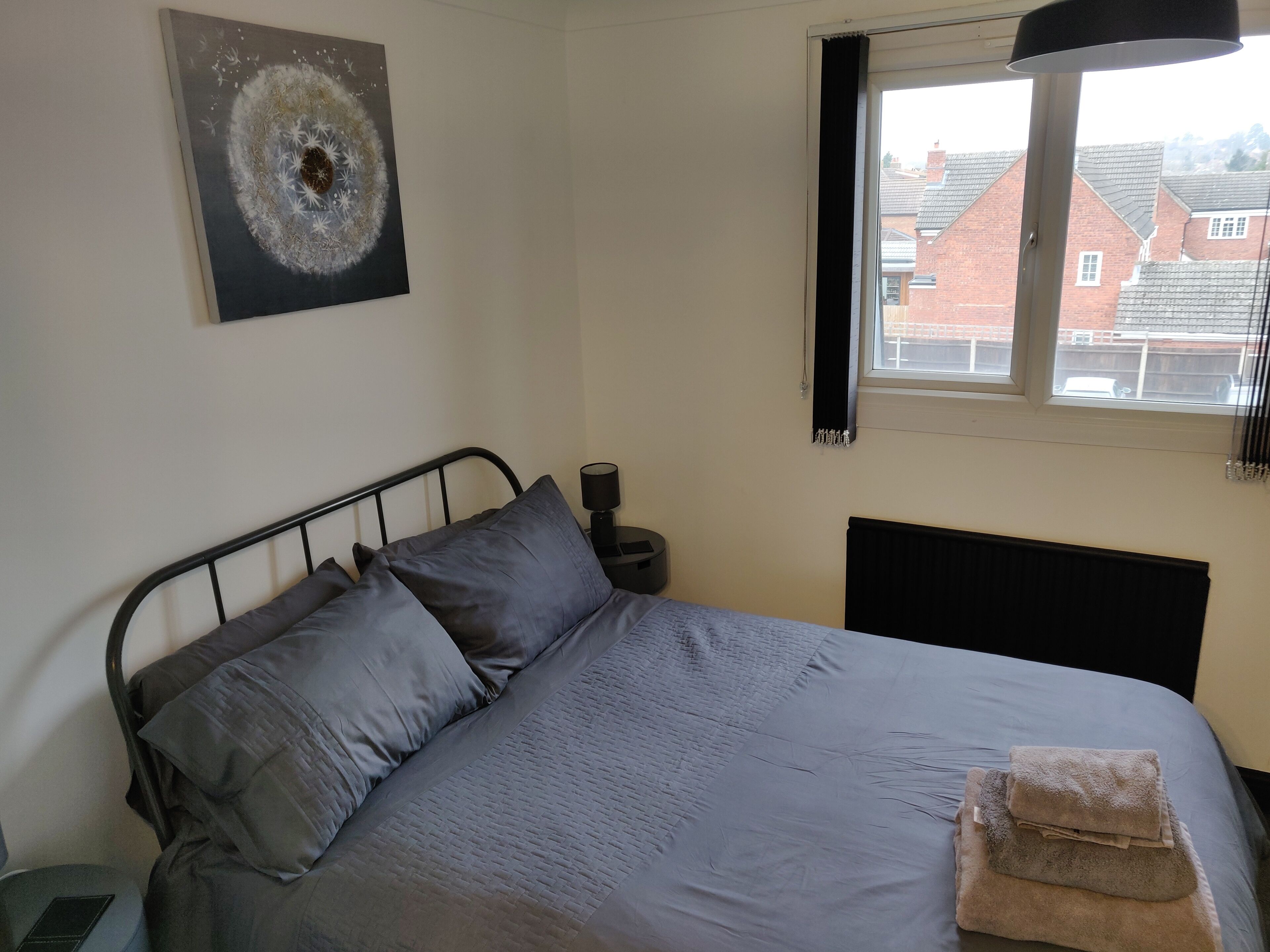 A bedroom with a bed, pillows, a bedside table with a lamp, a window, and a painting on the wall.